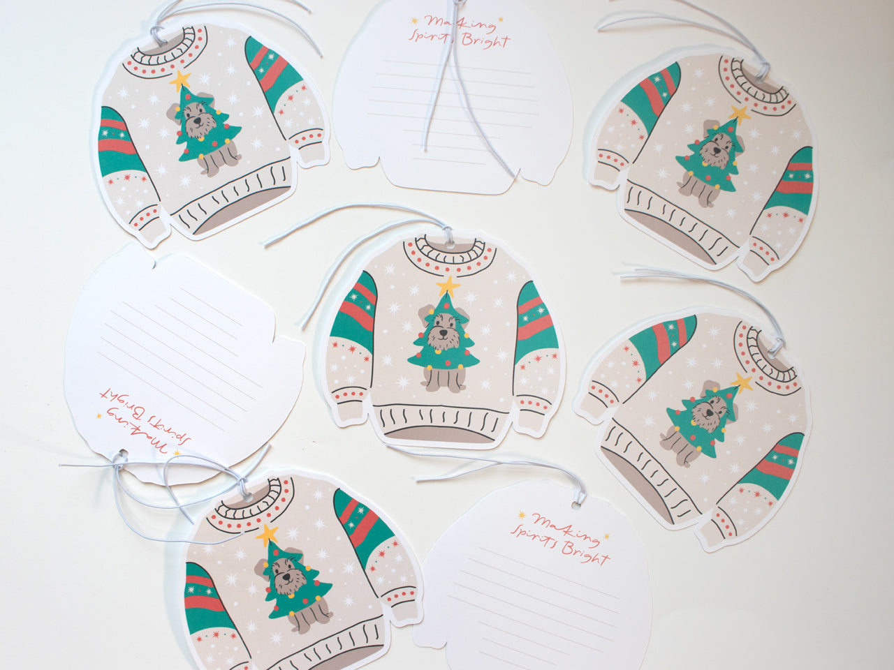 Display of multiple Christmas sweater-shaped gift tags spread across a surface. Each tag features a playful design of a schnauzer dog dressed as a Christmas tree, complete with ornaments and a yellow star, set against a beige sweater with festive red and green accents on the sleeves. Some tags are flipped over, revealing a space on the back for writing, along with the phrase “Making Spirits Bright” in cheerful red script. Each tag is equipped with a white string, ready to use on holiday gifts.