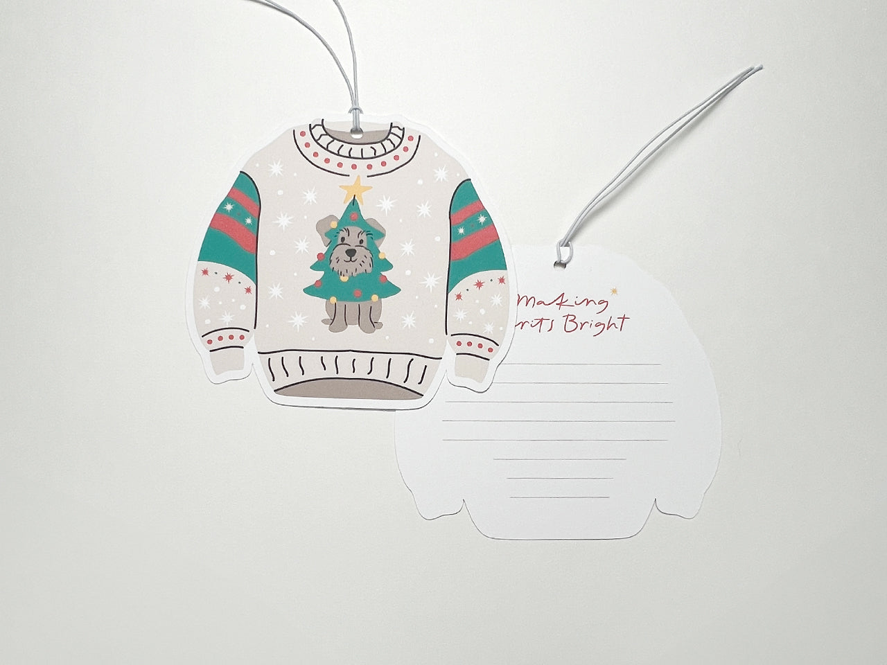 Two Christmas sweater-shaped gift tags placed side by side. The front of one tag features a schnauzer dog dressed as a Christmas tree, complete with ornaments and a yellow star, set against a beige sweater with festive red, green, and white accents on the sleeves. The second tag is flipped to show the back, which includes the phrase "Making Spirits Bright" in red script with a small yellow star, along with lined spaces for a personalized message.