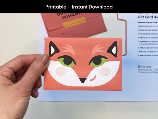 A person holding a completed fox printable gift card holder with bright green eyes and a smiling expression. This DIY fox envelope is a fun and colorful design for kids, teachers, or special occasions. Perfect for gifting money or gift cards. Shown with printed instructions in the background for easy assembly. Great for birthdays, thank you gifts, or last-minute printable gifts.