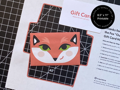 A flat lay of a DIY printable fox gift card holder template placed on a cutting mat, with the assembled envelope featuring a bold fox face design in the center. Ideal for 8.5 x 11 inch paper and perfect for digital downloads, craft projects, or last-minute gifts. Includes step-by-step instructions for printing, cutting, folding, and assembling at home. Great for handmade gifting.