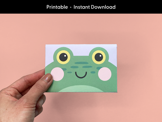 A hand holding a cute printable frog gift card holder against a pastel pink background, featuring a green frog face with yellow eyes and pink cheeks. The DIY envelope is designed for gifting and perfect for birthdays, celebrations, or appreciation gifts. The card holder is a completed version of the printable template, ideal for kids or fun themed occasions.