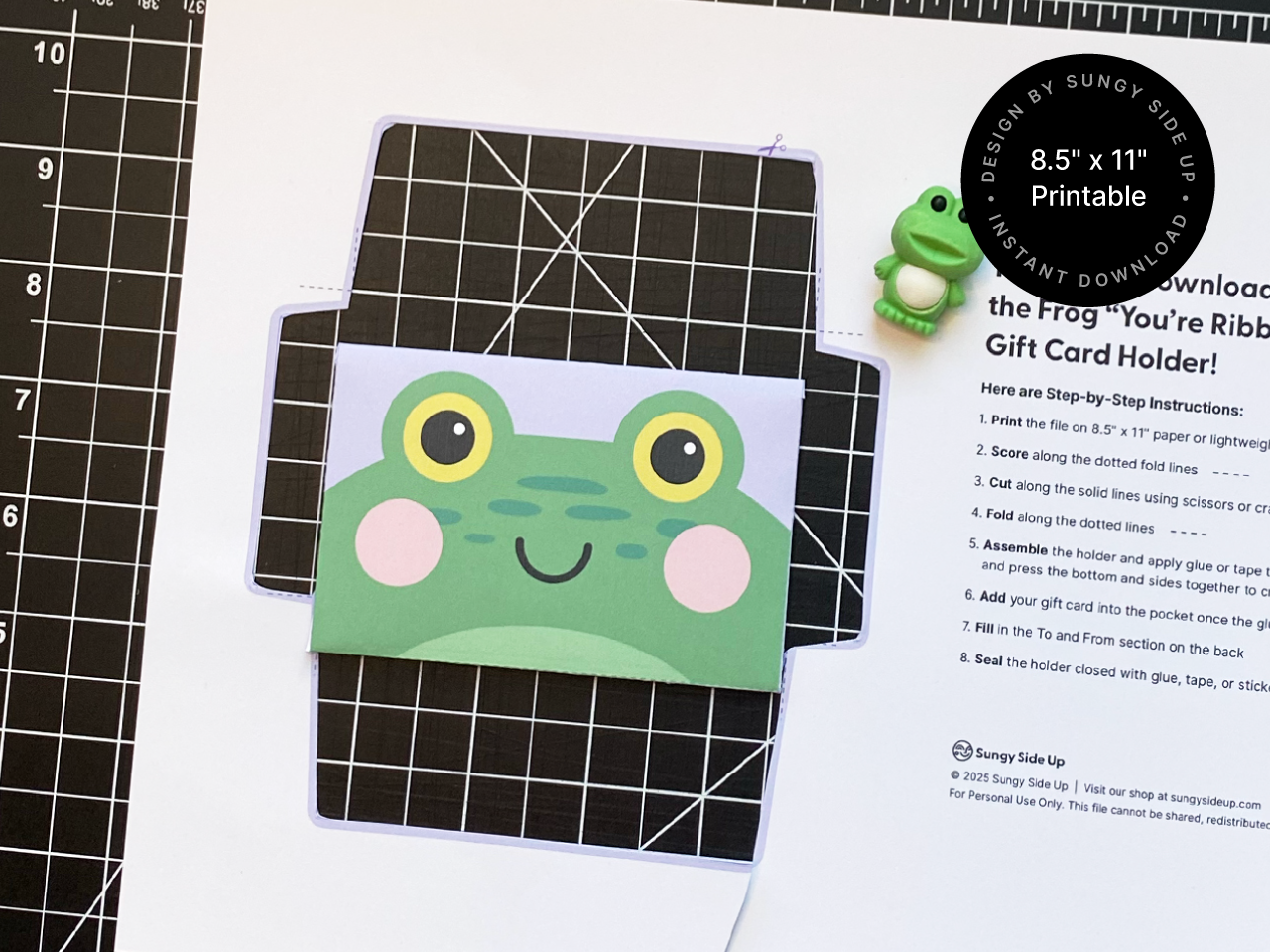 Printable frog gift card holder shown partially assembled on a cutting mat next to a small green frog figurine. The image showcases the colorful template being crafted, with a focus on the fun frog face design and step-by-step instructions for creating the envelope. This digital download is perfect for DIY celebrations, teacher gifts, birthdays, and party favors.
