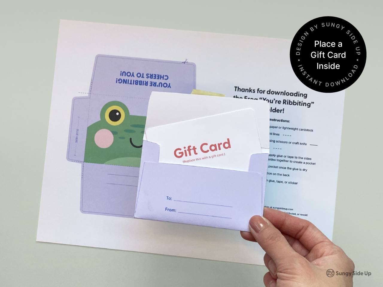 A hand holding an assembled printable frog gift card holder with a mock gift card inserted inside. The background shows the printed template and step-by-step assembly instructions. The design features a cheerful green frog face, making it perfect for birthdays, thank you gifts, or general celebrations. Ideal for DIY gifting and printable craft projects.