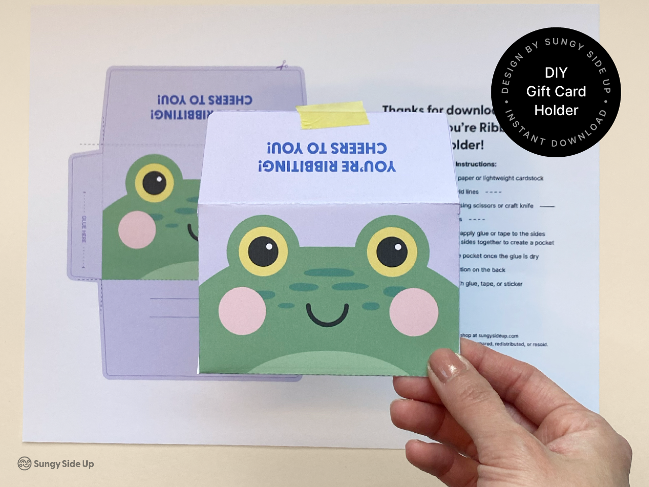 Close-up of a completed frog gift card holder being held above the printed template sheet. The assembled card holder showcases a playful frog design with bright yellow eyes and pink cheeks, ideal for printable DIY gifts. The background includes clear step-by-step instructions, making this a great option for handmade celebration or appreciation gifts.