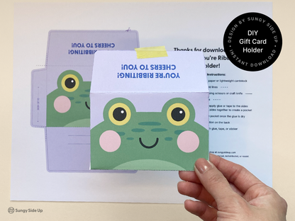 Close-up of a completed frog gift card holder being held above the printed template sheet. The assembled card holder showcases a playful frog design with bright yellow eyes and pink cheeks, ideal for printable DIY gifts. The background includes clear step-by-step instructions, making this a great option for handmade celebration or appreciation gifts.