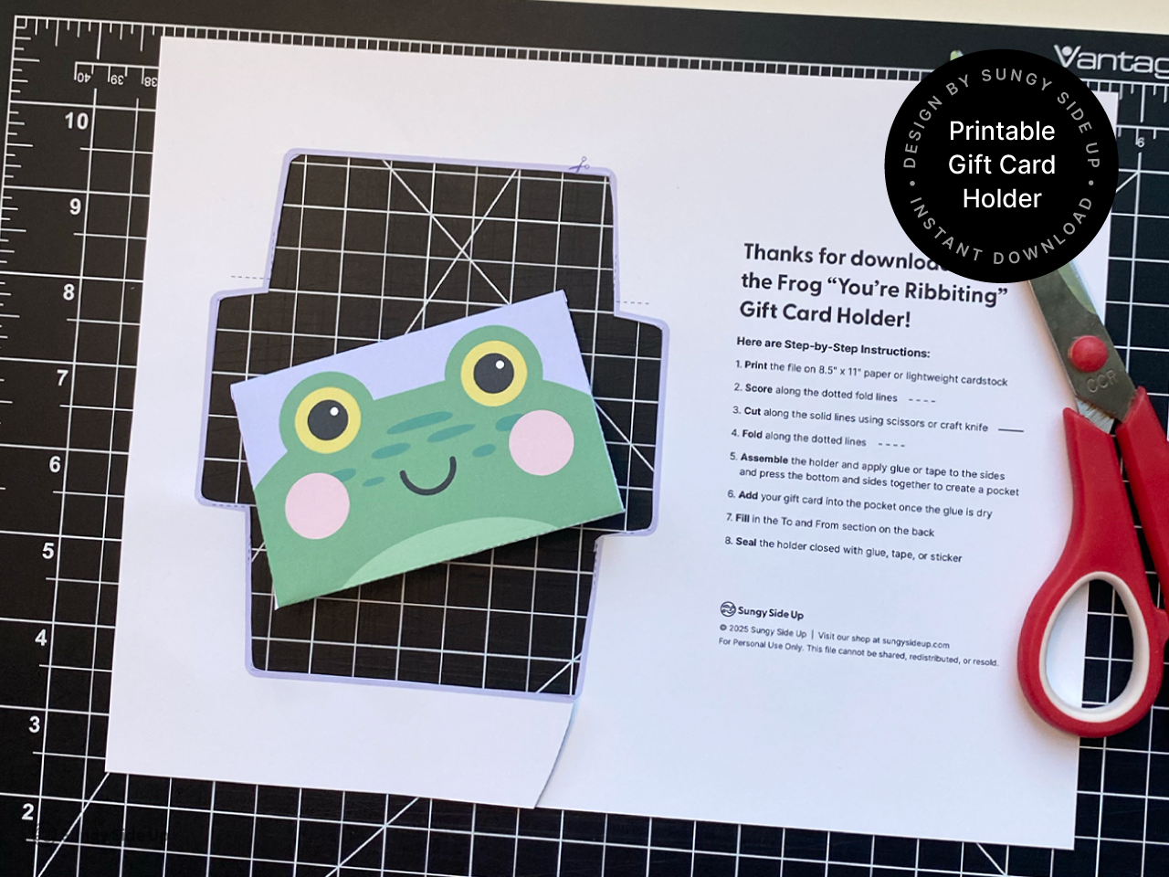 A printable frog-themed gift card holder partially cut and assembled on a black crafting mat with red scissors beside it. The image highlights the crafting process, showing the colorful frog face design and included step-by-step instructions. This digital download is perfect for creating fun and personal DIY gift card envelopes for birthdays, thank yous, or special occasions.