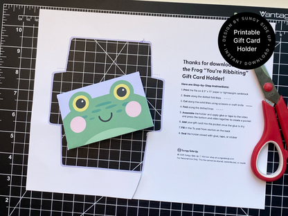 A printable frog-themed gift card holder partially cut and assembled on a black crafting mat with red scissors beside it. The image highlights the crafting process, showing the colorful frog face design and included step-by-step instructions. This digital download is perfect for creating fun and personal DIY gift card envelopes for birthdays, thank yous, or special occasions.