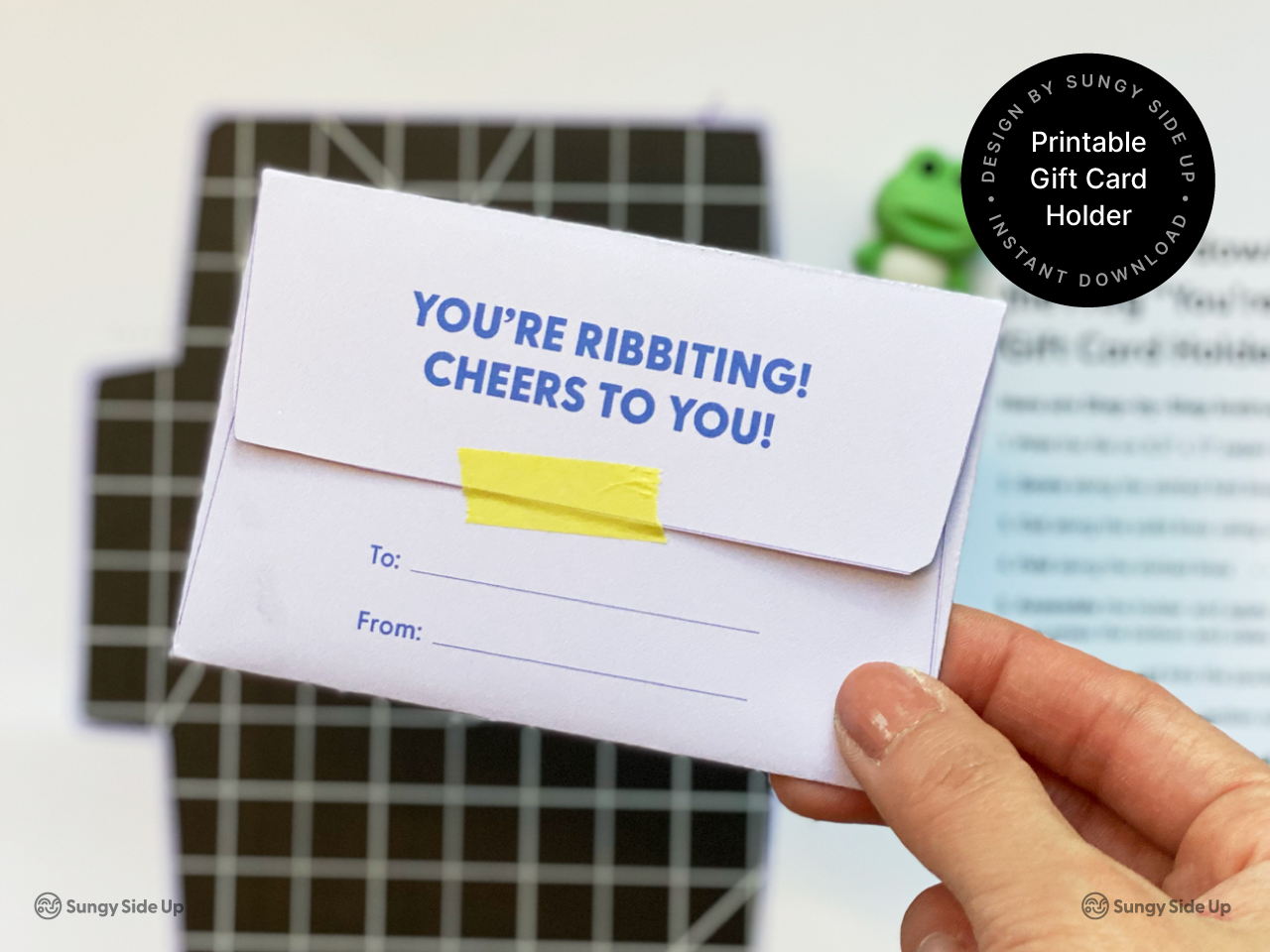 A hand holding the back view of an assembled frog-themed printable gift card holder. The envelope features a cheerful message and a section to write recipient and sender names. The card is sealed with yellow tape and placed over a cutting mat and printed instruction sheet, perfect for DIY gifting and handmade crafts.