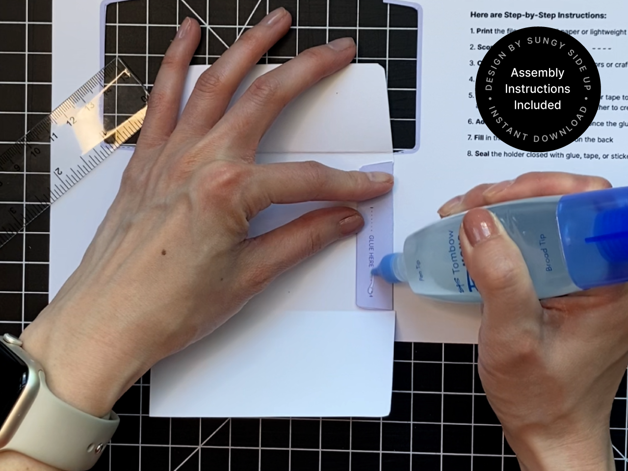 Close-up of hands applying glue to the flap of a frog-themed printable gift card holder using a precision tip glue bottle. The template is laid on a black cutting mat with a ruler nearby, showing the crafting process involved in assembling the envelope. Ideal for DIY gift giving, birthdays, or appreciation gifts.