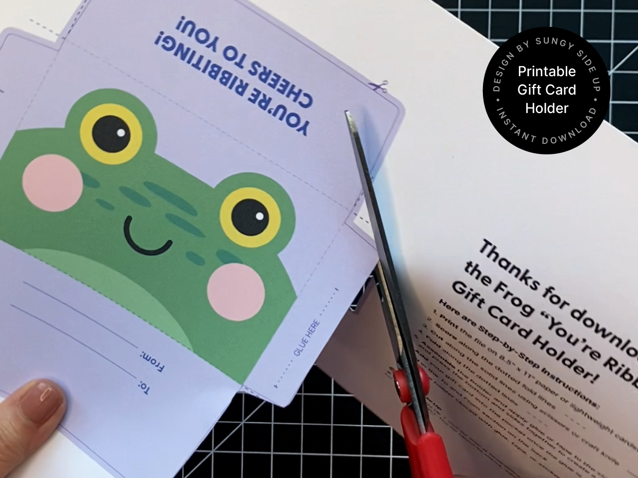 A hand cutting out the frog printable gift card holder along the solid lines using scissors. The template features a green frog face with rosy cheeks and a to/from section on the back. The background includes the full instruction page, highlighting the hands-on crafting nature of the digital download.