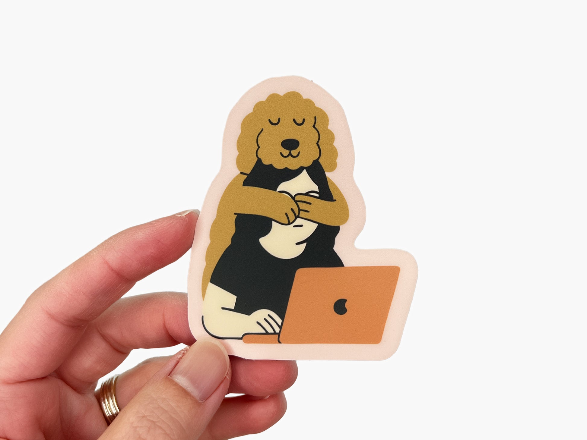 Hand holding a matte die cut sticker showing an illustration of a golden doodle hugging its human from behind while the person works on a laptop. The sticker has a soft pink border and warm neutral colors on a white background.