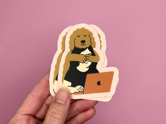Hand holding a small stack of matte die cut stickers against a pink background. Each sticker shows an illustration of a golden doodle hugging its human from behind while the person works on a laptop. The sticker has a soft pink border and warm neutral colors.