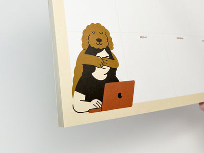 Close-up of a weekly planner notepad showing a calm dog illustration with curly fur hugging a person seated at a desk and working on a laptop. The illustration appears in the corner of a clean Monday–Sunday planner layout, printed on paper with a soft, neutral color palette.