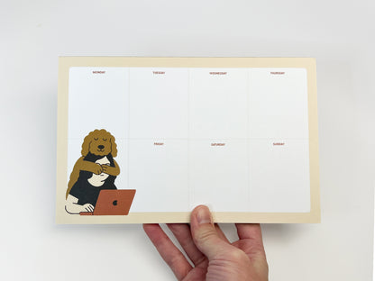 A hand holds an 8.5 x 5.5 inch weekly planner notepad with a clean Monday–Sunday layout. In the lower left corner, a calm dog illustration with curly fur hugs a person working on a laptop. The notepad features a soft, neutral color palette and ample blank space for weekly planning.