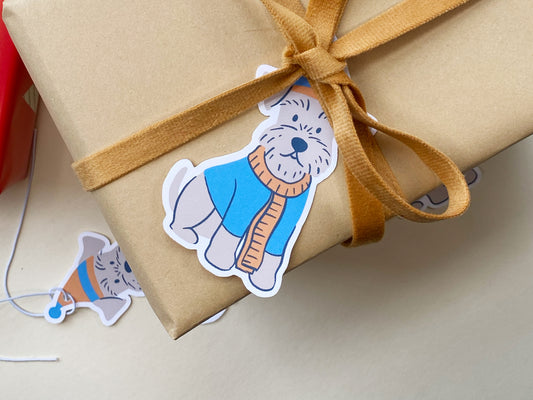 Handmade Schnauzer gift tag tied to a kraft-wrapped present with a mustard yellow velvet ribbon. The illustrated Schnauzer dog features a cozy blue and orange winter hat and scarf, adding a festive, pet-friendly touch to holiday gift wrapping.