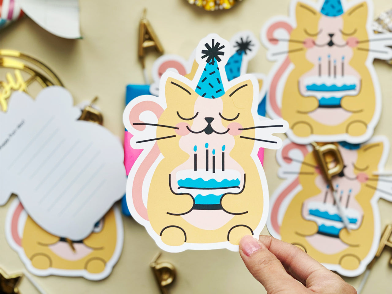 A close-up of a hand holding a cat-shaped birthday card featuring an illustrated yellow cat with a pink tail, wearing a blue party hat, and holding a birthday cake with lit candles. The background displays other cards, envelopes, and gold decoration.