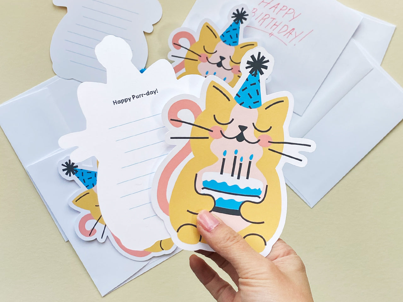 A hand holds a cat-shaped birthday card with a cute yellow cat illustration wearing a blue party hat and holding a birthday cake. The back of the card has lined space with the phrase Happy Purr-day printed at the top.