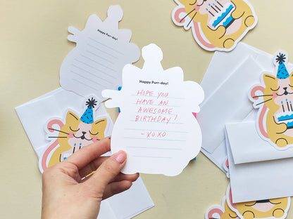 A close-up of the inside of a cat-shaped birthday card with the printed phrase Happy Purr-day and handwritten text in red ink. The background includes matching illustrated cards of a cat holding a cake and white envelopes.