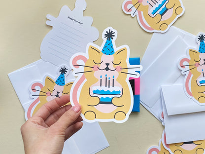 A hand holds a cat-shaped birthday card featuring a cute illustrated yellow cat wearing a blue party hat and holding a blue cake with lit candles. The card has a playful design and reads Happy Purr-day! on the back with lined space for writing.