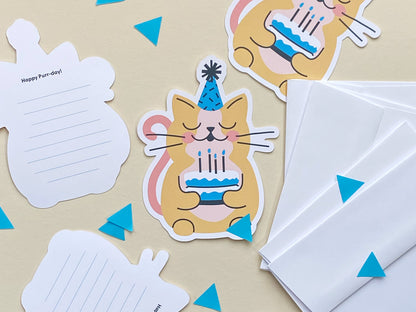 A flat lay display of cat-themed birthday cards featuring a cute yellow cat wearing a blue party hat and holding a blue birthday cake. The cards are arranged with white envelopes and show some of the back of cards which are lined for messages.