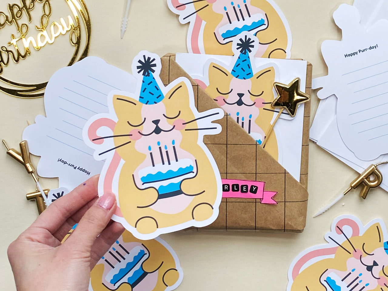 A hand holds a cat-shaped birthday card featuring an illustrated yellow cat wearing a blue party hat and holding a blue birthday cake. The background includes other flat cards, white envelopes, and gold birthday decorations like a cake topper.