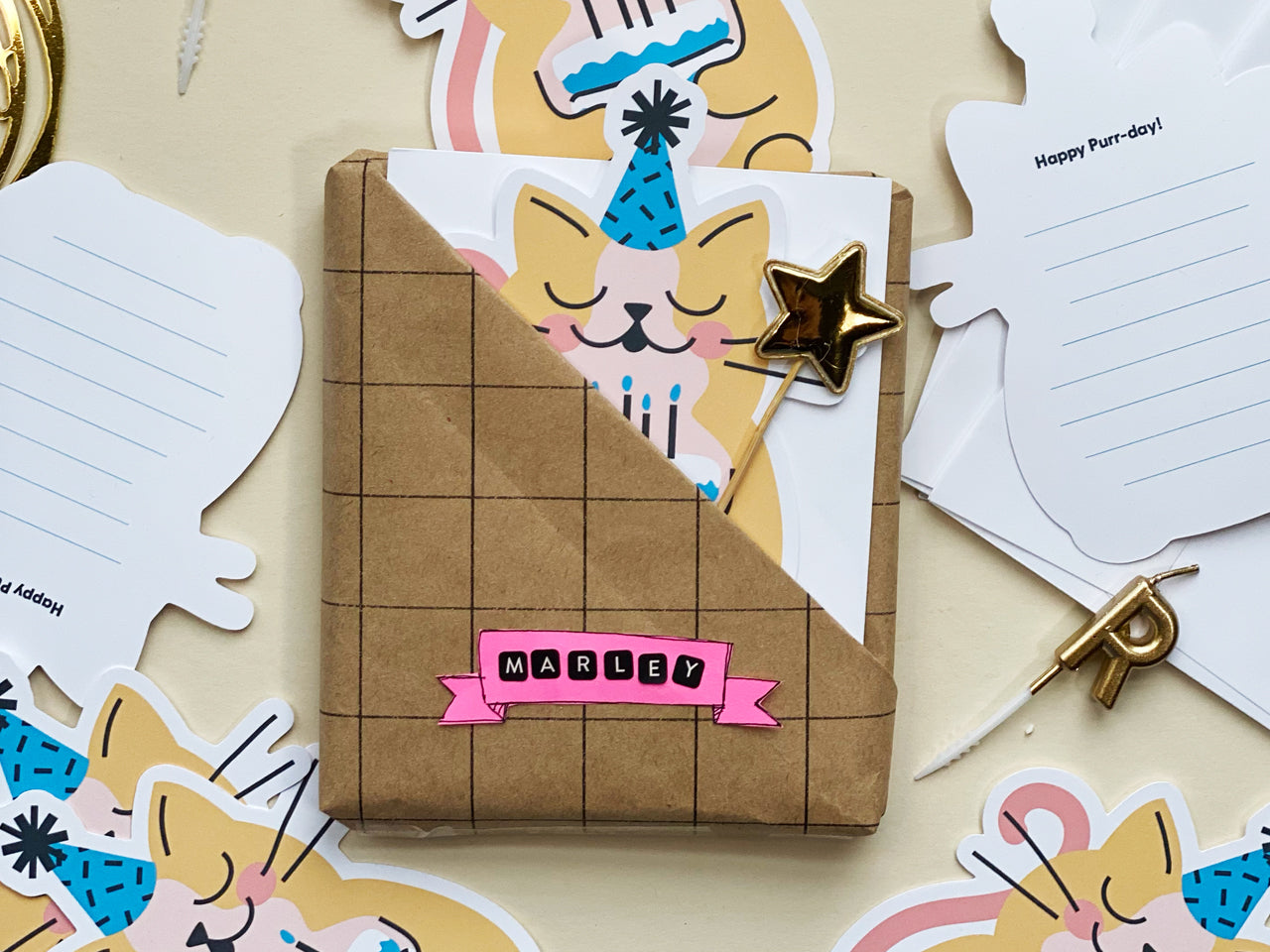 A gift wrapped in grid-patterned brown paper with a pink label displayed alongside cat-themed birthday cards. The cards feature a yellow cat wearing a blue party hat and holding a birthday cake. A gold star topper decorates the package.