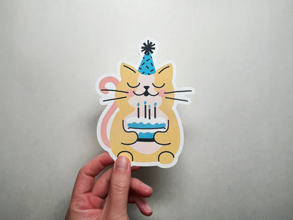 A close-up of a hand holding a cat-shaped birthday flat note card featuring an illustration of a smiling yellow cat wearing a blue party hat and holding a birthday cake with lit candles.