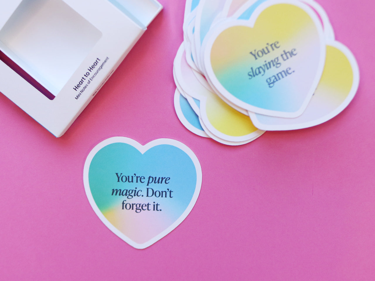 A close-up of gradient heart-shaped encouragement cards with phrases such as You’re pure magic. Don’t forget it. The cards are spread out on a pink surface beside their packaging.