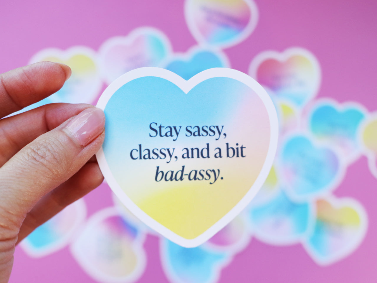 A hand holding a heart-shaped encouragement note card with a gradient design and sassy quotes like the phrase Stay sassy, classy, and a bit bad-assy. Several similar cards are blurred in the background.