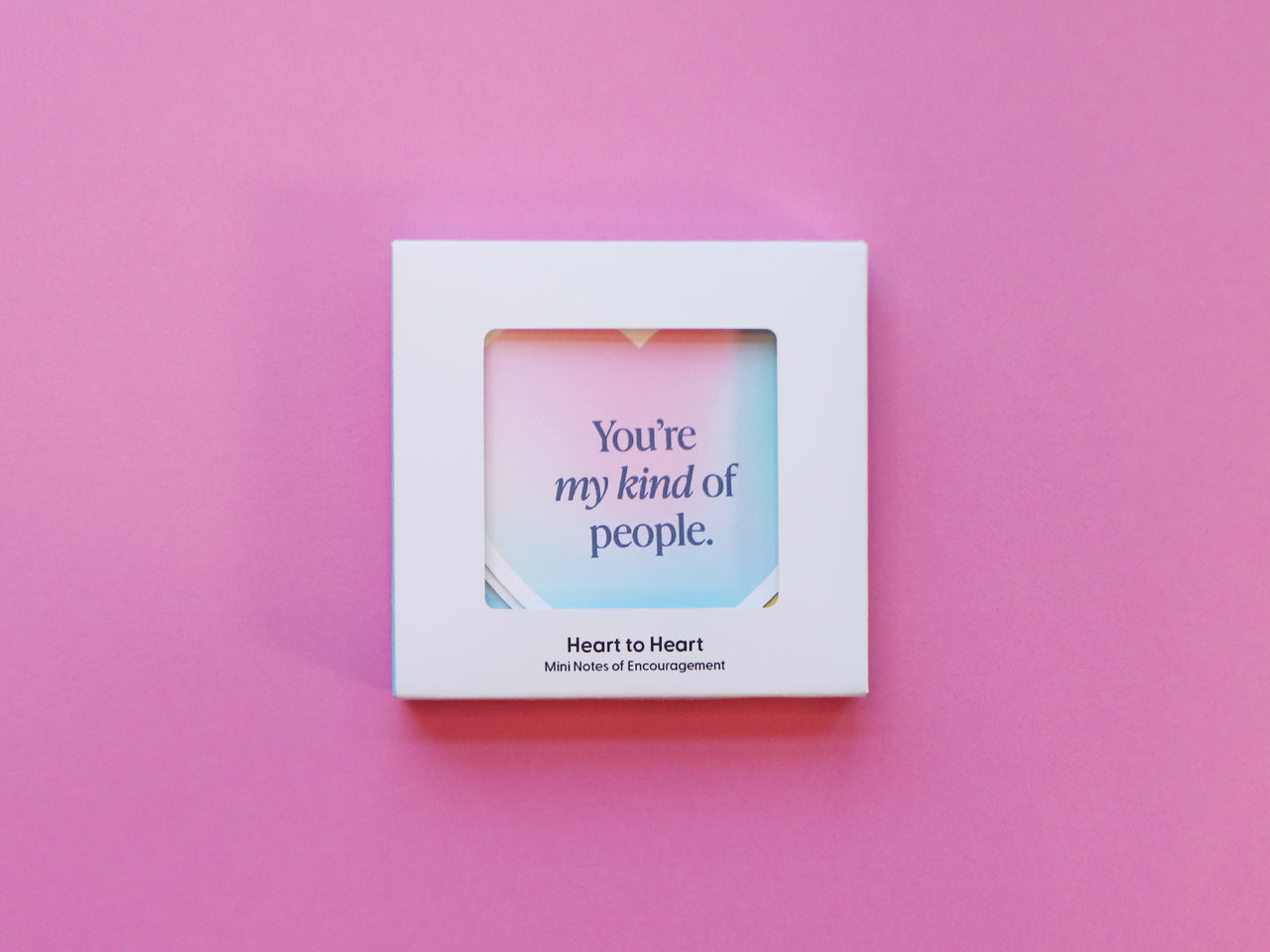 A closed white packaging box on a pink background with a gradient-colored heart-shaped card visible through a window. The visible card says, You’re my kind of people. This is aset of motivational mini note cards that would be giftable for friends.