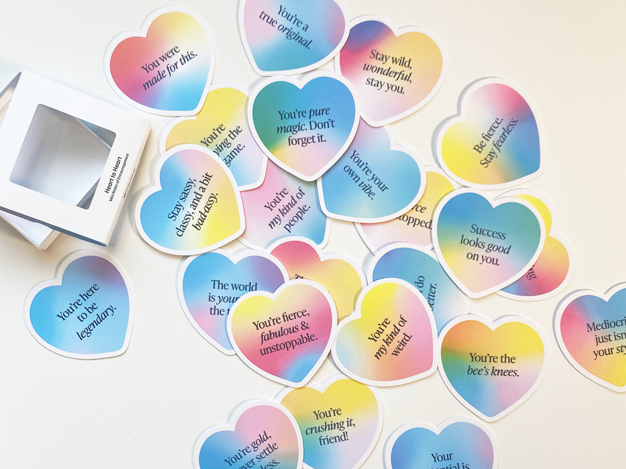 A spread of heart-shaped encouragement note cards with gradient pastel designs featuring inspiring phrases. An open white packaging box labeled sits beside the colorful cards, which are perfect for gifting, sharing positivity, or personal stationery.