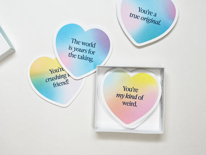 An open box of heart-shaped gradient note cards on a white surface. Visible messages include You’re my kind of weird, The world is yours for the taking, and “ou’re crushing it, friend! showcasing colorful, motivational mini notes arranged casually.