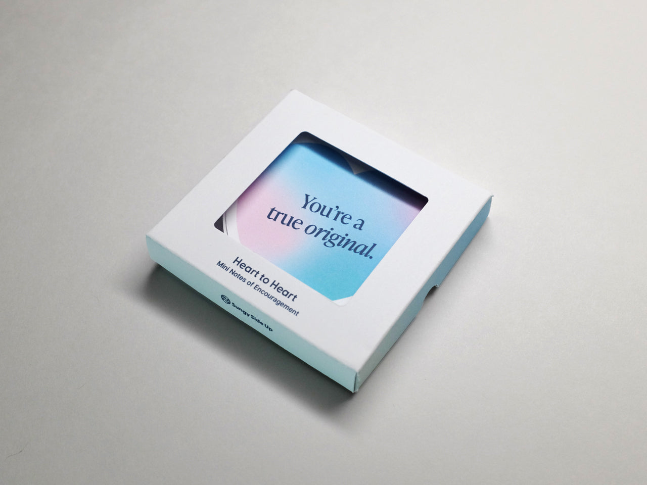 A closed white box of Heart to Heart: Mini Notes of Encouragement with a heart-shaped card visible through the window. The blue and pink gradient card displays the phrase, You’re a true original, against a clean, neutral background.