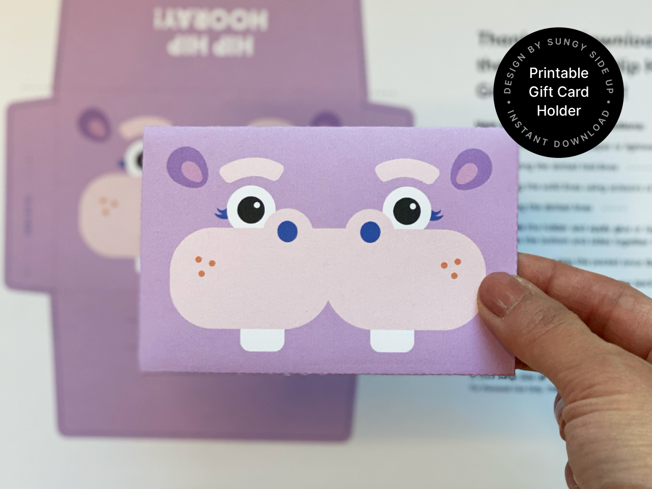 Close-up of a completed hippo gift card envelope held in front of a printed template sheet. This printable product is a digital download for making fun and easy gift card holders at home. Perfect for birthdays, classroom rewards, and kids celebrations. The cute hippo design adds charm to any gift and is ideal for those looking for DIY paper crafts or handmade card alternatives.