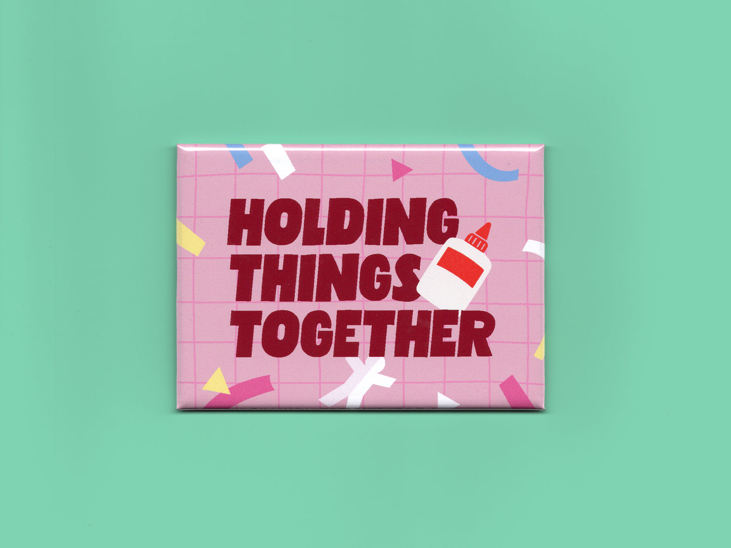 Glossy rectangular magnet with a pink grid background, colorful confetti shapes, and bold dark red text that reads “Holding Things Together.” A small glue bottle illustration overlaps the text. The magnet is displayed against a mint green background.