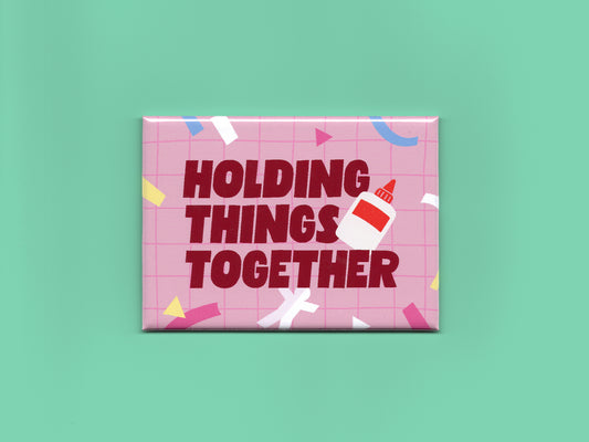 Glossy rectangular magnet with a pink grid background, colorful confetti shapes, and bold dark red text that reads “Holding Things Together.” A small glue bottle illustration overlaps the text. The magnet is displayed against a mint green background.