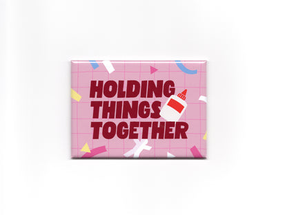 Rectangular pink magnet with a light pink grid pattern, colorful confetti accents, and bold dark red text that reads “Holding Things Together.” A small glue bottle illustration overlaps the text on the right side.