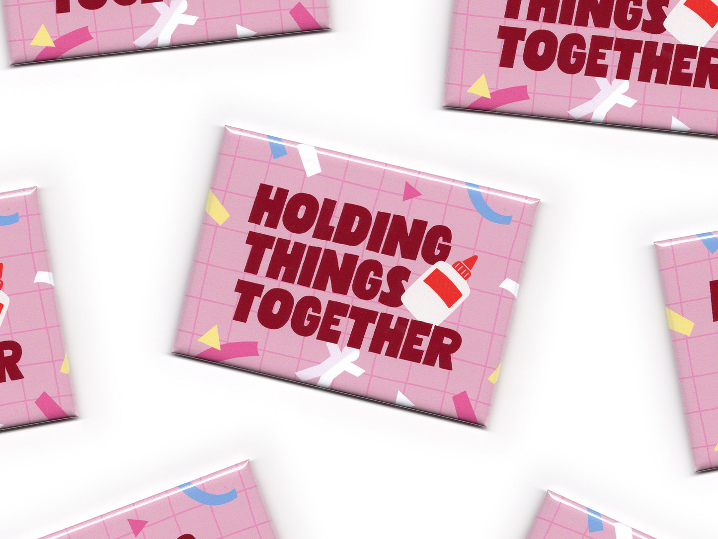 Multiple pink “Holding Things Together” magnets scattered across a white background. The magnets feature a pink grid pattern, colorful paper cut accents, bold dark red text, and a small glue bottle illustration.