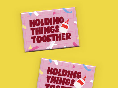 Two pink “Holding Things Together” magnets displayed on a bright yellow background. The magnets feature a pink grid pattern, colorful paper cut out scrap accents, bold dark red lettering, and a small glue bottle illustration.