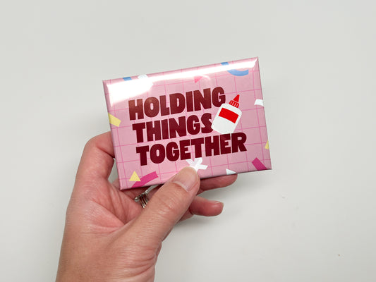 Hand holding a glossy rectangular magnet with a pink grid background, colorful confetti accents, and bold dark red text that reads “Holding Things Together.” A small glue bottle illustration appears beside the text.