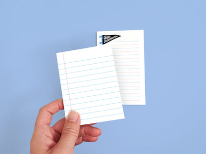 Hand holding a mini lined notepad with a Chaos Coordinator notepad in the background, showing the size and paper design of the notepad set.