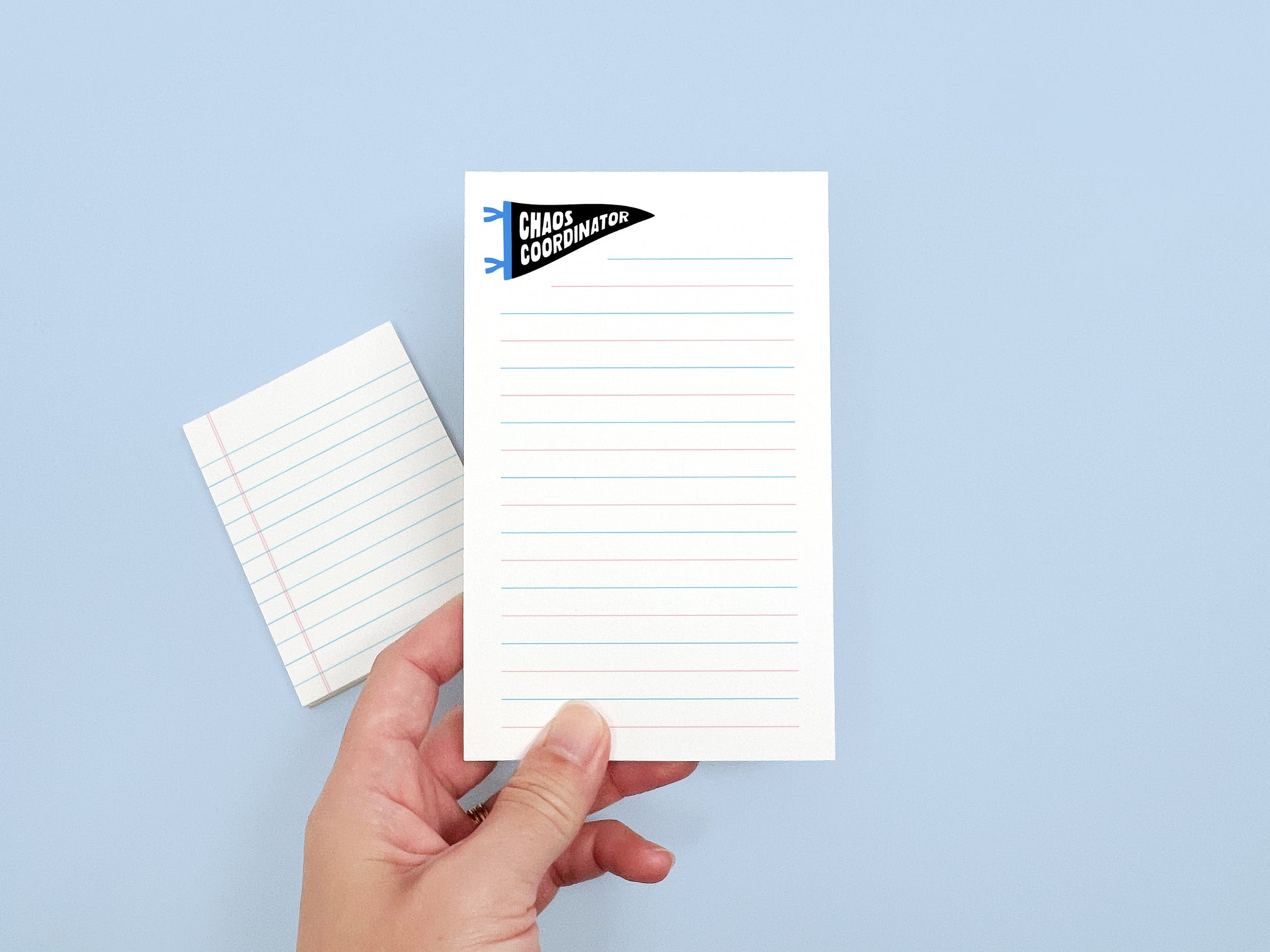 Hand holding a Chaos Coordinator lined notepad with a matching mini notepad in the background, showing the size and design of the notepad set.