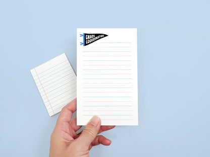 Hand holding a Chaos Coordinator lined notepad with a matching mini notepad in the background, showing the size and design of the notepad set.