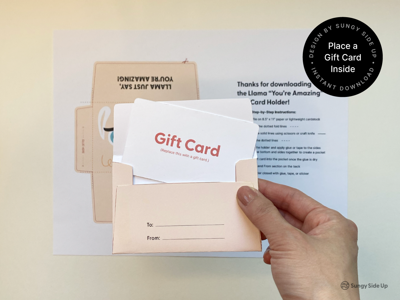 Hand inserting a mock gift card into a finished printable llama envelope. The background shows the original printed template and step-by-step assembly instructions. This image demonstrates how to use the DIY llama gift card holder, making it ideal for product listings targeting keywords like printable gift card envelope, kids birthday gift, teacher appreciation idea, and animal themed card holder for instant download.