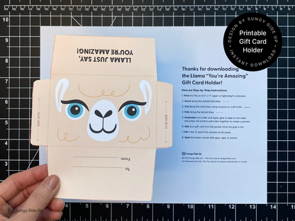 Close-up of a hand holding a printed llama gift card holder template above a black cutting mat. The image shows the template prior to assembly with printed folding lines and instructional sheet in the background. Great for promoting printable gift card envelopes, DIY craft templates, kids party ideas, and teacher appreciation gifts. This is a digital download perfect for last-minute or creative gifting with a handmade feel.
