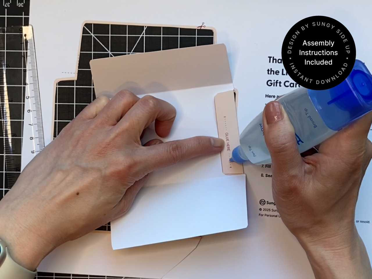 Close-up of hands applying glue to the edge of a printed llama gift card holder template during the assembly process. A ruler and cutting mat are partially visible in the background, emphasizing the DIY nature of the project. This image supports keywords like how to assemble printable gift card holders, DIY animal envelope, llama gift craft, and printable gift wrap idea for small gifts. Great for crafters and gift givers.