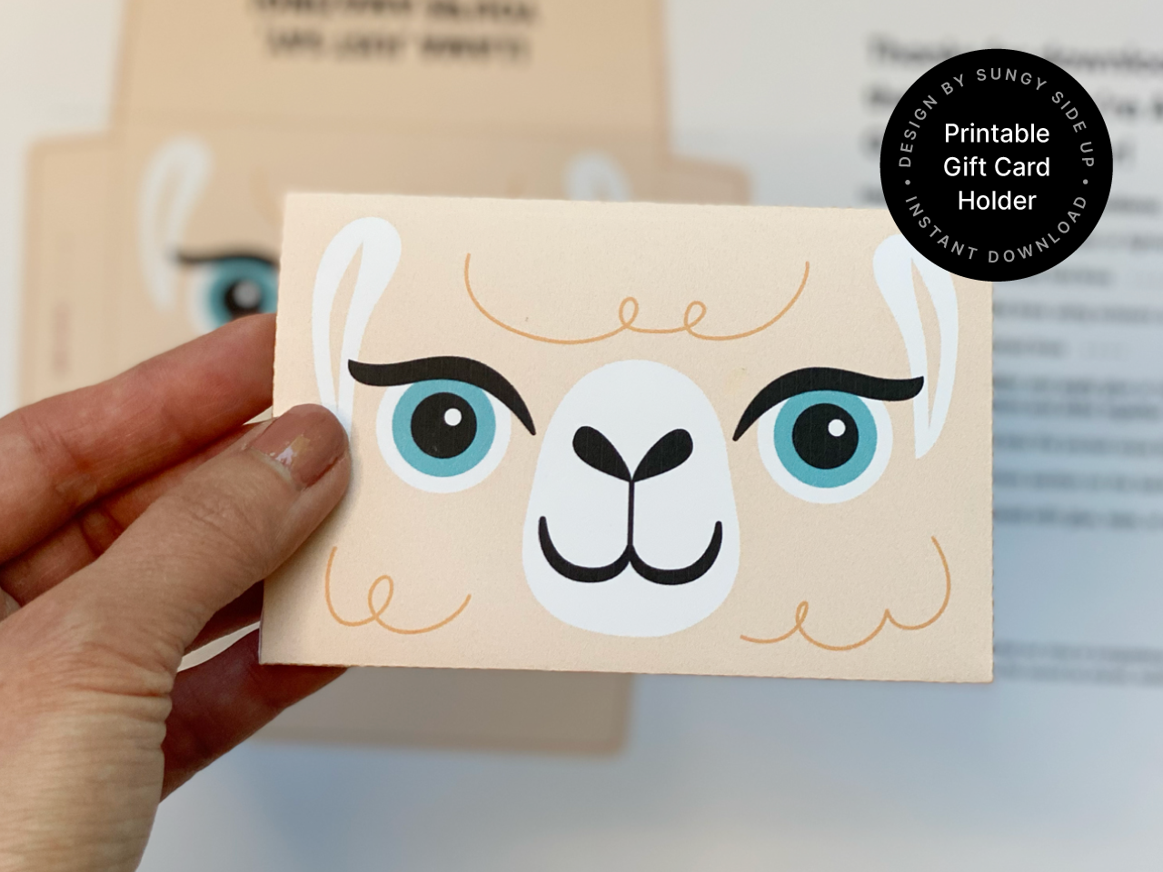 Close-up of a hand holding a completed llama gift card envelope in front of a blurred background with the printed template. The llama design is clearly visible, making it the focal point. Perfect for search terms like printable llama gift card holder, cute DIY gift wrap idea, instant download envelope for gift cards, or animal themed gift packaging. Great for birthdays, thank you gifts, and party favors.