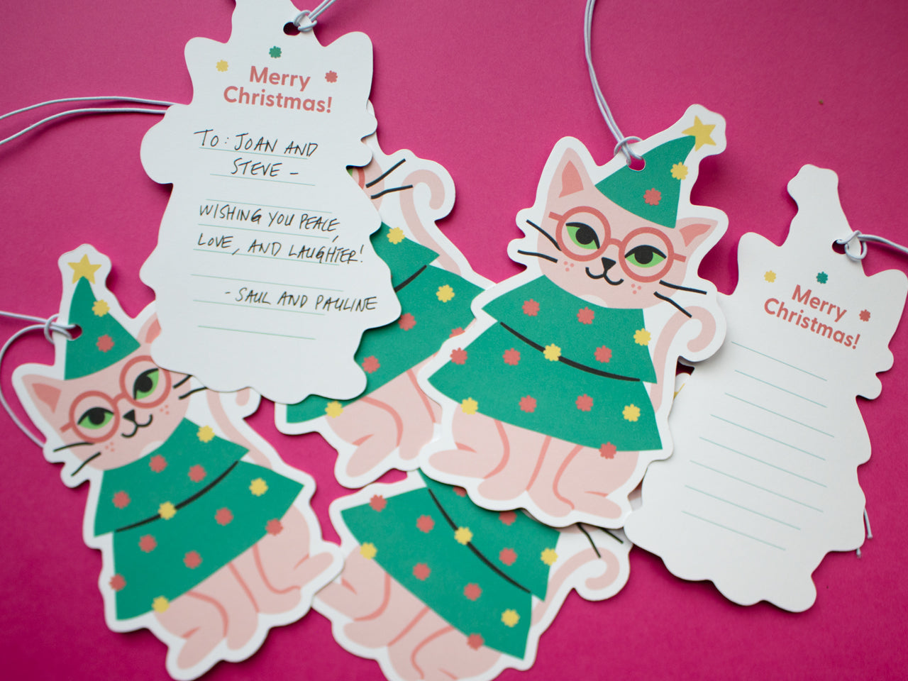 Multiple gift tags with a cat wearing glasses wearing a Christmas tree outfit spread across a magenta surface. The gift tags feature the cat illustration on the front with "Merry Christmas" on the back with lines to write a special message.