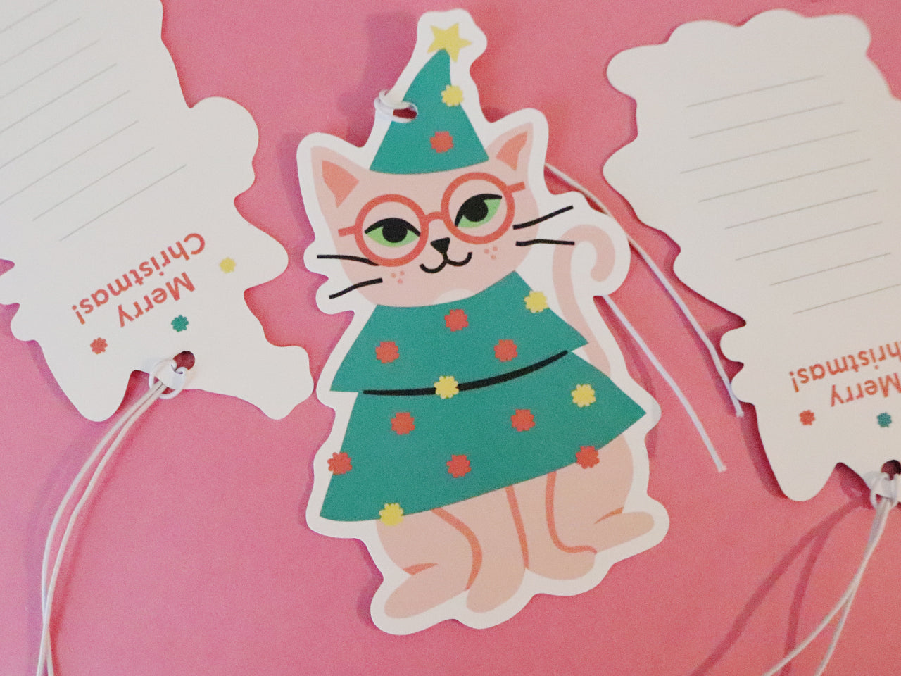 Holiday gift tags shaped like a cat dressed as a Christmas tree. The whimsical cat illustration features pink fur, round glasses, and a Christmas tree outfit decorated with colorful ornaments and topped with a star. The back of the tags is visible on the left and right sides of the image, where the words "Merry Christmas!" are printed in festive red text, along with some space for writing a personalized message.