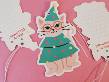 Holiday gift tags shaped like a cat dressed as a Christmas tree. The whimsical cat illustration features pink fur, round glasses, and a Christmas tree outfit decorated with colorful ornaments and topped with a star. The back of the tags is visible on the left and right sides of the image, where the words "Merry Christmas!" are printed in festive red text, along with some space for writing a personalized message.